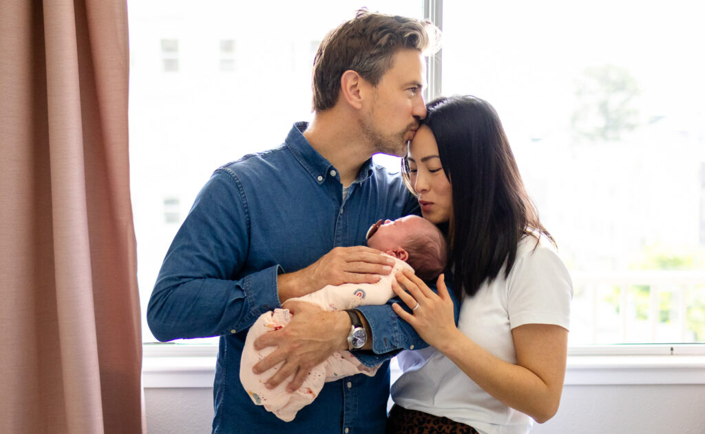 Father gently kisses mother’s forehead while both cradle their newborn during In Home vs Studio Newborn Photography comparison