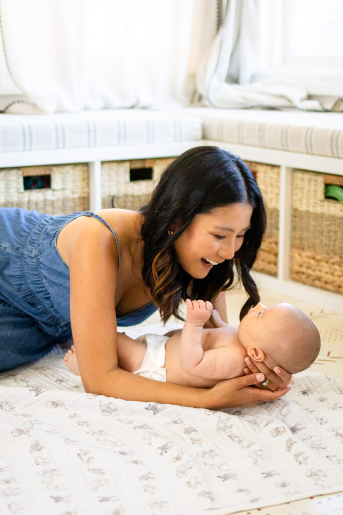 Mother lying on the floor smiling down at her diapered newborn during In Home vs Studio Newborn Photography comparison