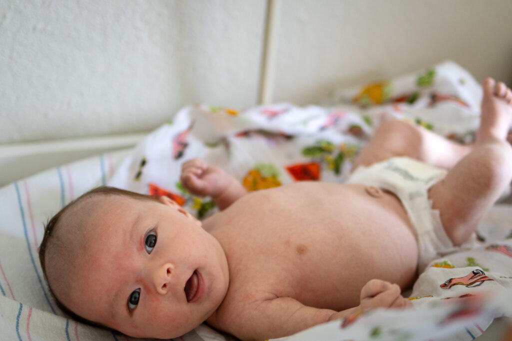 Newborn lying on a bed wearing only a diaper, looking toward the camera in soft natural light.