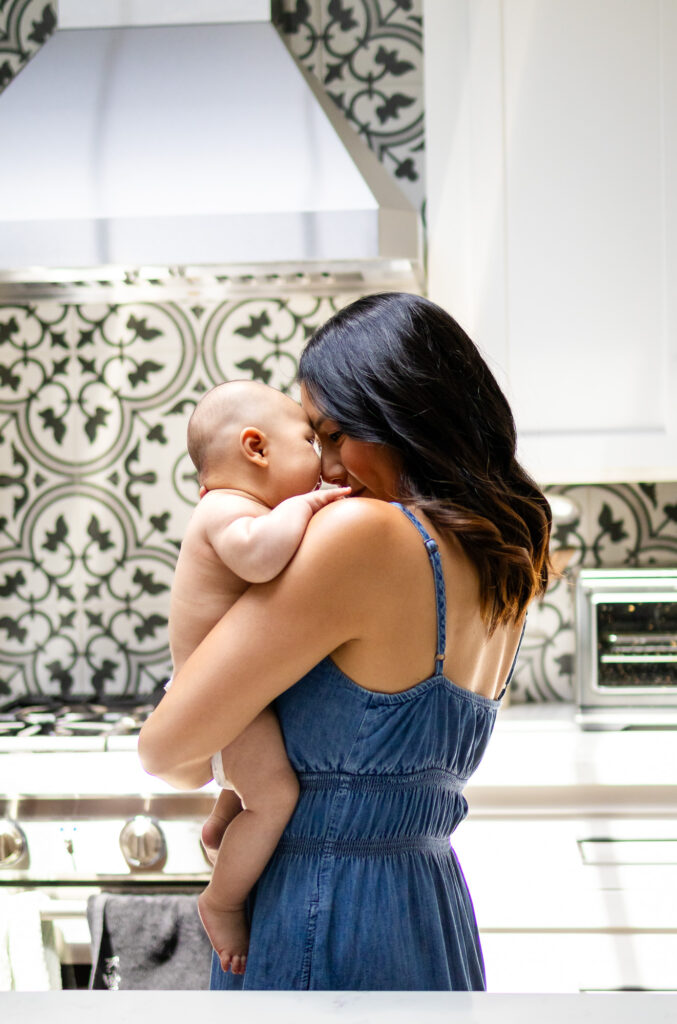 Mother standing in a kitchen holding her baby close against her shoulder near patterned tile backsplash.