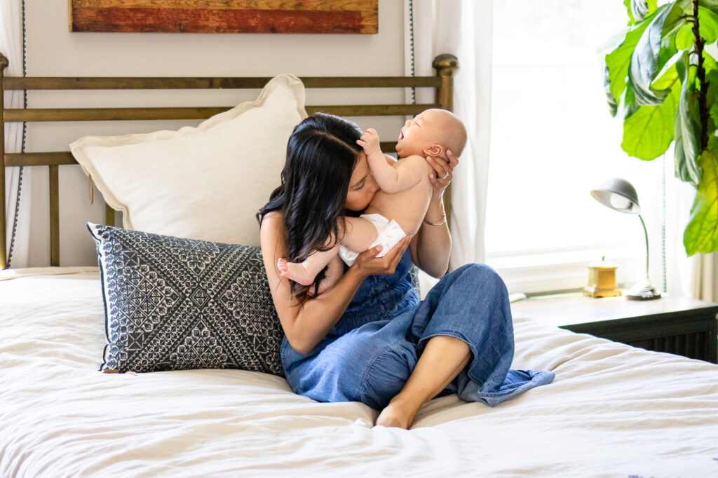 Mother sitting on a bed kissing her baby during In Home vs Studio Newborn Photography comparison