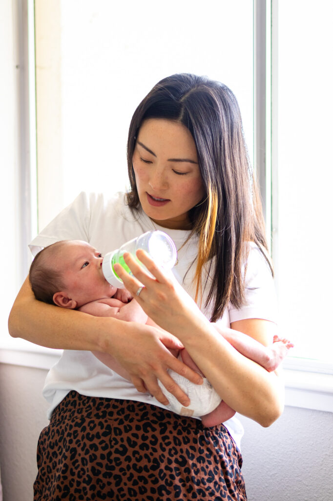 A mother stands near a bright window holding and bottle feeding her newborn baby, natural light softly illuminating both of them.