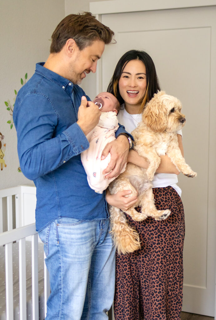 Parents stand together in their nursery holding their newborn baby and their small tan dog, a sweet example of Including Pets in Newborn Photos at Home.