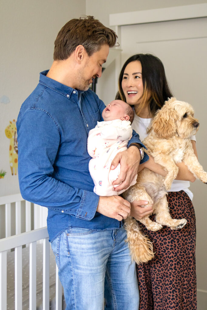 A father cradles his newborn while the mother holds their small dog, both parents smiling warmly at their baby during an in home newborn session.