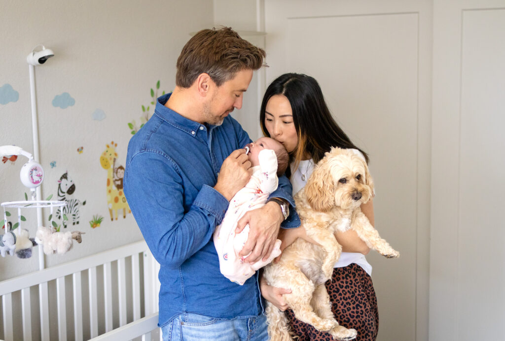A mother kisses her newborn baby on the forehead while holding their small dog, capturing Including Pets in Newborn Photos at Home in a natural and heartfelt way.