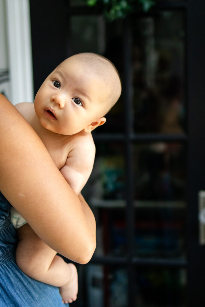 Baby resting upright in a parent’s arms near a window, looking outward with soft natural light highlighting their face.