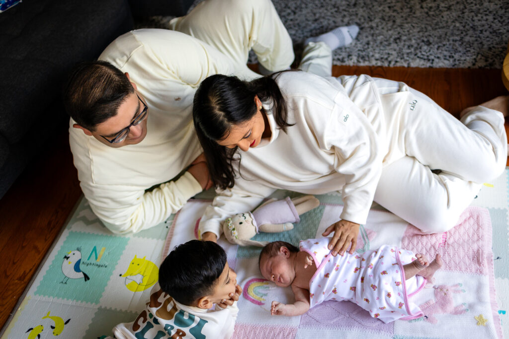 Parents and toddler lying on a play mat beside their swaddled newborn, all looking down at the baby in a relaxed family moment.