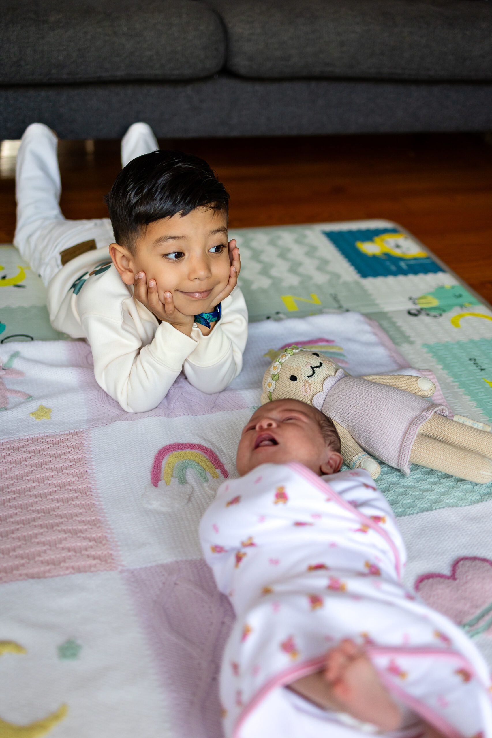 Toddler lying on their stomach beside their swaddled newborn sibling, resting their chin in their hands and smiling.