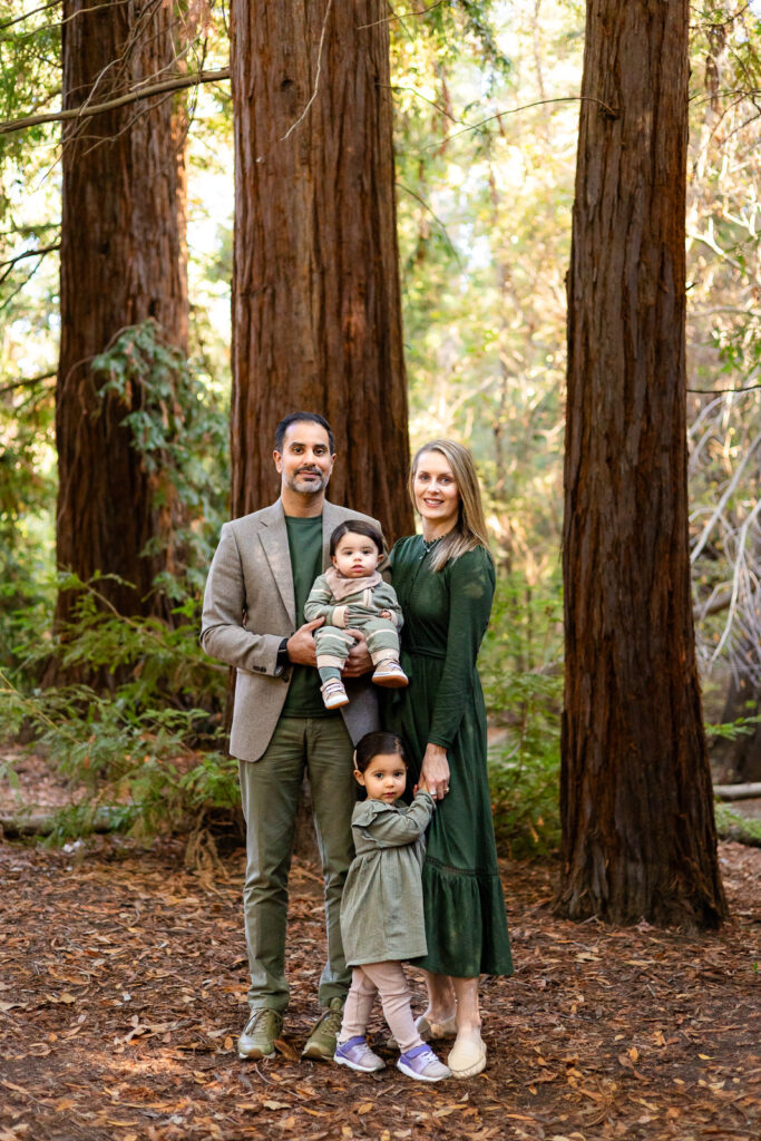 A family of four stands together among tall redwood trees, parents holding their baby while their toddler stands close, surrounded by soft forest light