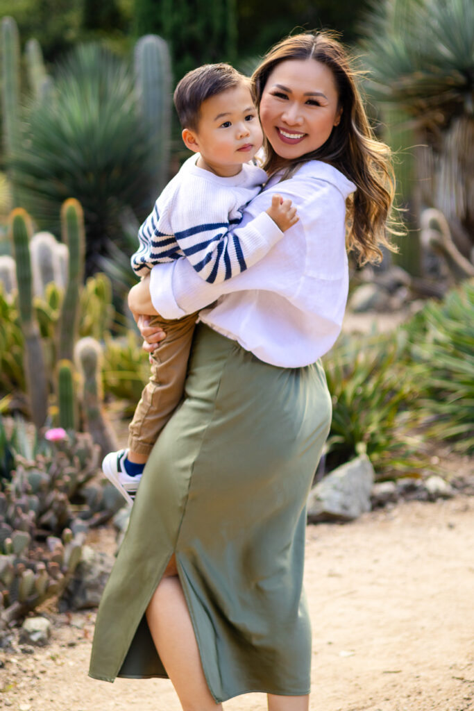 A mother smiles while holding her young son in her arms, standing among cacti and desert plants
