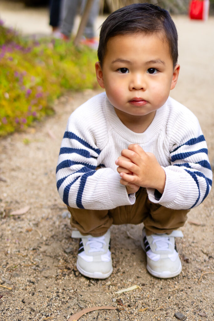 A toddler crouches on a garden path looking directly at the camera, hands clasped together during a quiet moment at a scenic photo location in Palo Alto.