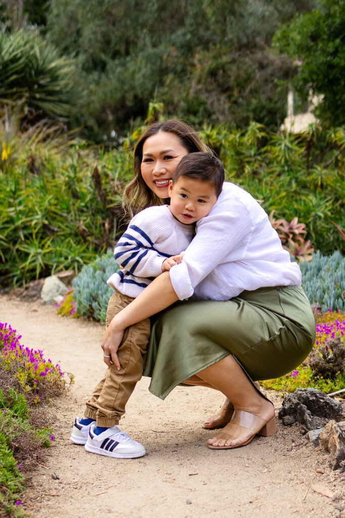 A mother crouches to hug her toddler close while smiling, framed by lush plants at a family friendly photo spot in Palo Alto.