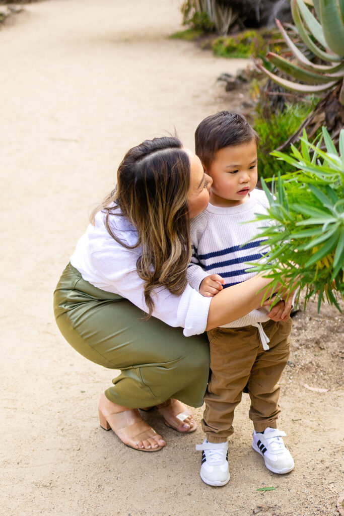 A mother bends down to kiss her toddler’s cheek as he looks at nearby plants, captured at a natural light photo park in Palo Alto.