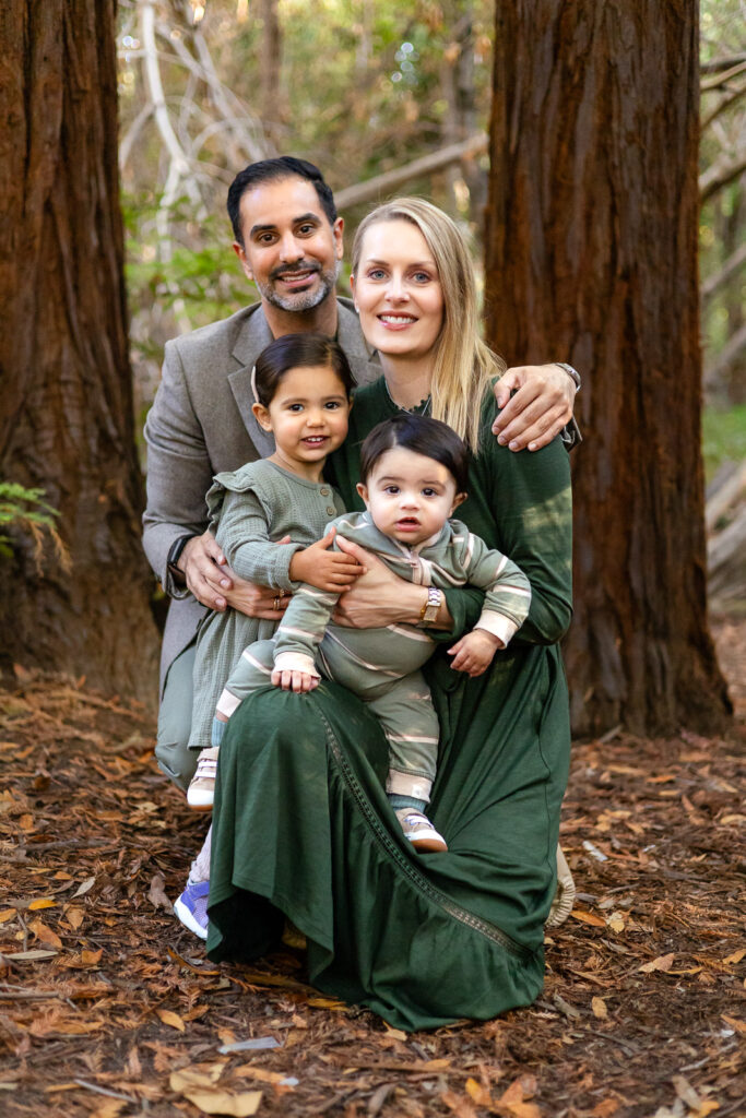 A family sits close together on the forest floor with their two children, sharing a quiet moment among the trees at one of the Palo Alto family photo locations.