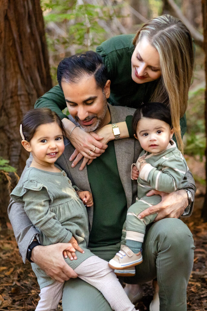 A mother and father crouch together with their toddler and baby, surrounded by tall trees at an outdoor photography park in Palo Alto.