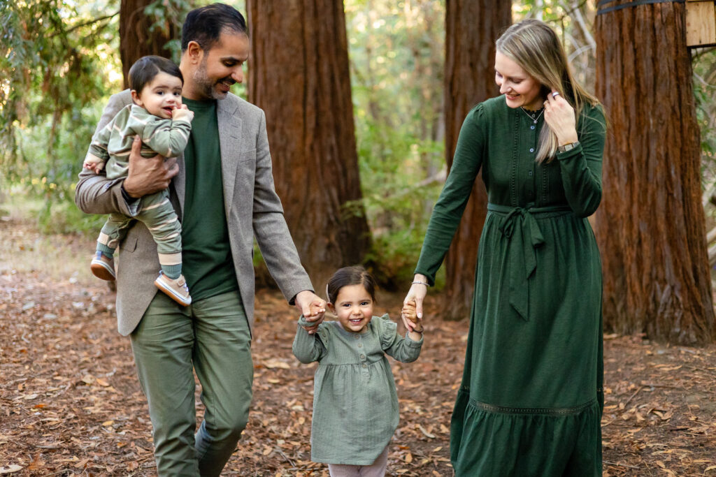 Parents walk hand in hand with their toddler while the father carries their baby, enjoying a quiet stroll through one of the beautiful photo forrest around Palo Alto.