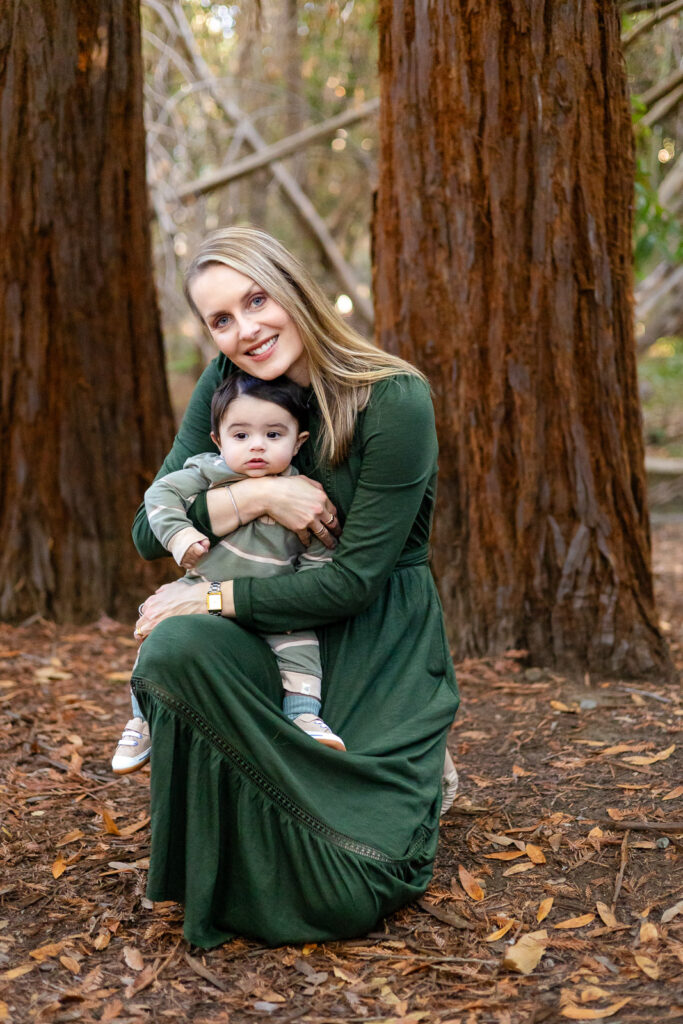 A mother holds her baby while sitting on the forest floor, smiling gently in soft light