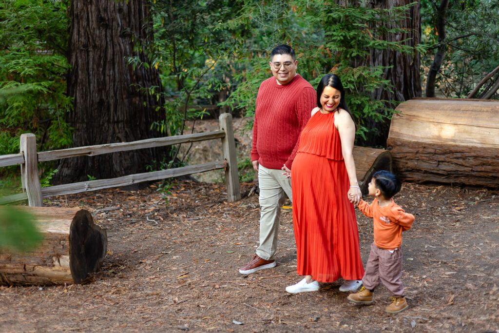 Expectant mother in a flowing orange dress walking through a wooded park while holding her toddler’s hand, her partner smiling beside them during lifestyle maternity photography in the Bay Area.