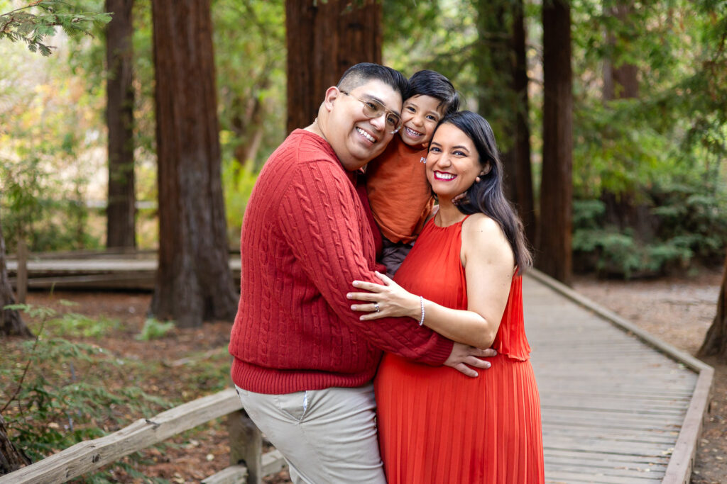 Parents standing on a wooden path in a redwood grove, hugging their smiling toddler between them during maternity photography with toddlers in the Bay Area.