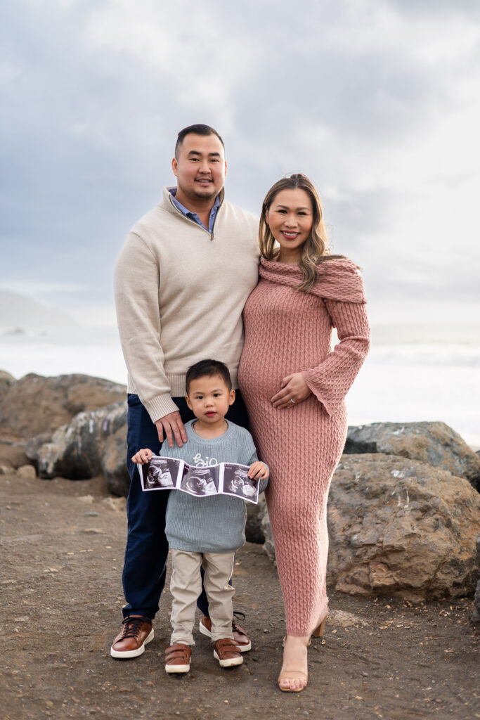 Expectant parents standing on rocky shoreline with their young son holding ultrasound photos, ocean waves behind them during Pregnancy Photography in the Bay Area.