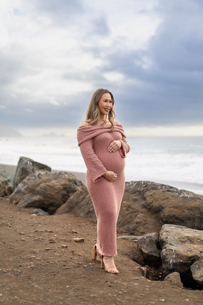 Expectant mother in a knit dress standing on seaside rocks, gently cradling her belly while looking toward the ocean during relaxed pregnancy photos on the Peninsula.