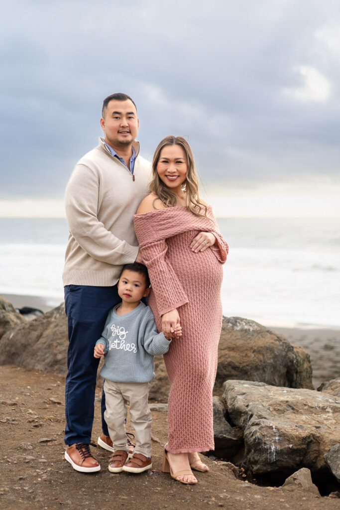 Parents standing together on rocky beach with their toddler holding hands, mother resting one hand on her belly during Pregnancy Photography in the Bay Area.