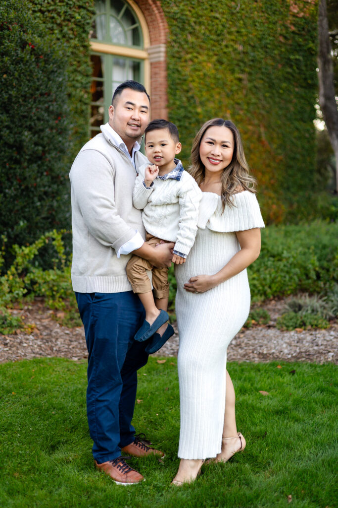 Expectant parents holding their toddler in front of an ivy covered brick building, smiling softly during Peninsula maternity photography.