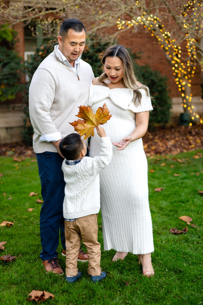 Parents smiling as their toddler hands them a large autumn leaf on a lawn covered with fallen leaves, capturing heartfelt pregnancy photos near San Mateo.