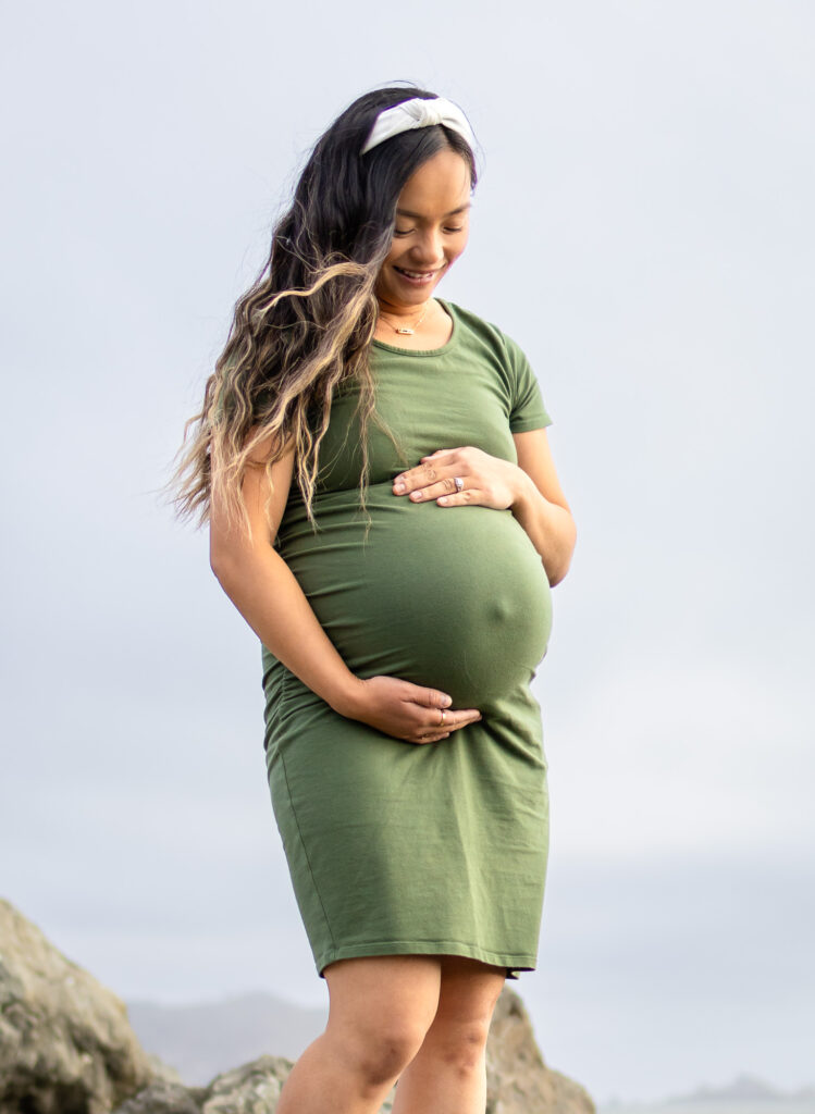 Expectant mother in a green dress standing on coastal rocks, looking down at her baby bump and smiling during Bay Area pregnancy photography.