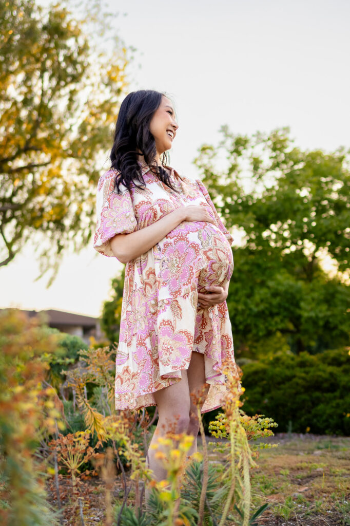 Expectant mother standing outdoors among greenery at sunset, cradling her belly and smiling softly during Peninsula maternity photography.