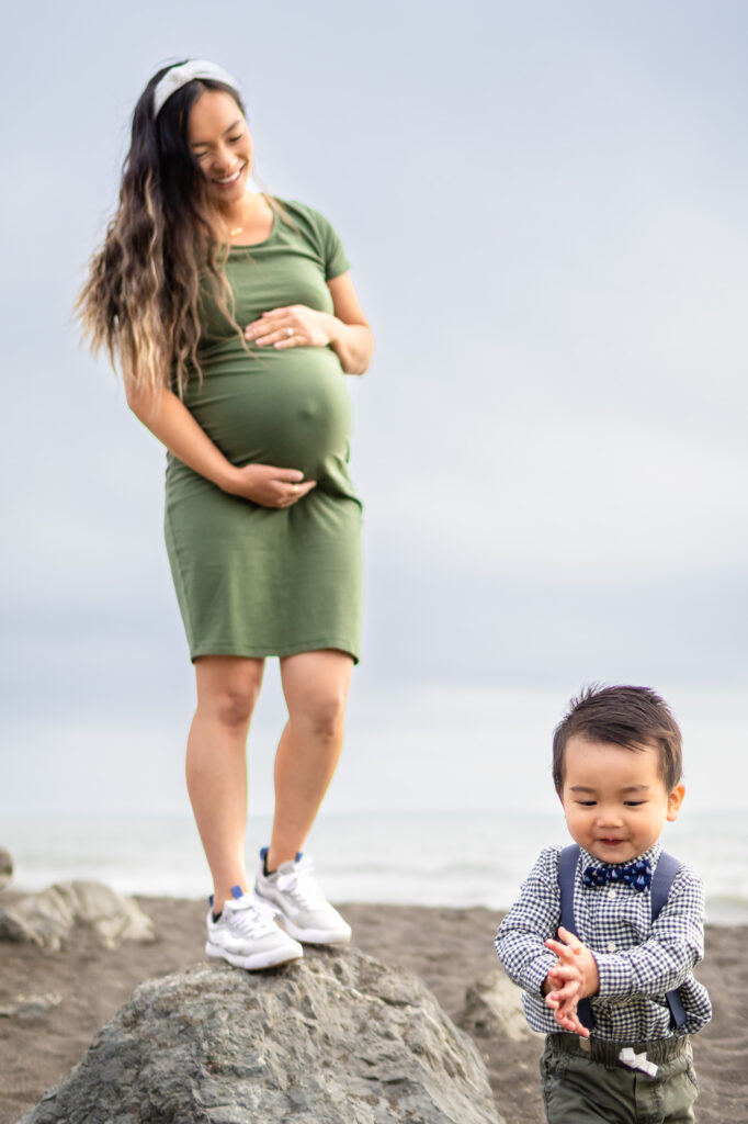 Pregnant mother in a green dress standing on coastal rocks while her toddler walks in front of her on the beach during relaxed pregnancy photos on the Peninsula.