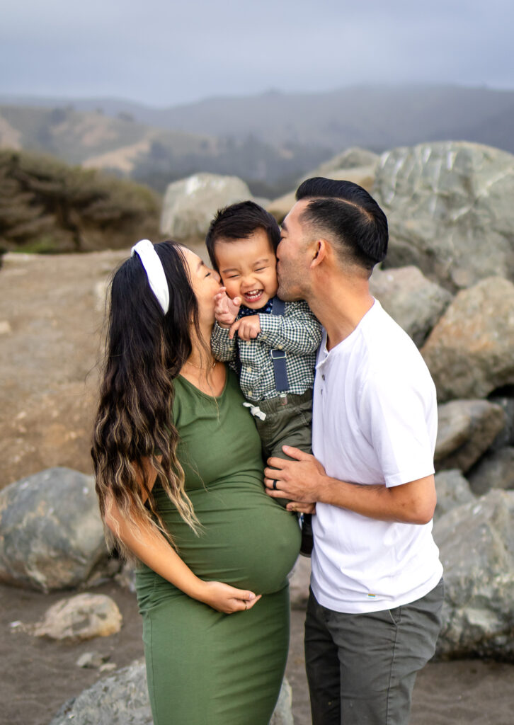 Parents kissing their smiling toddler’s cheeks while the mother cradles her baby bump on a rocky beach during Bay Area maternity photography.
