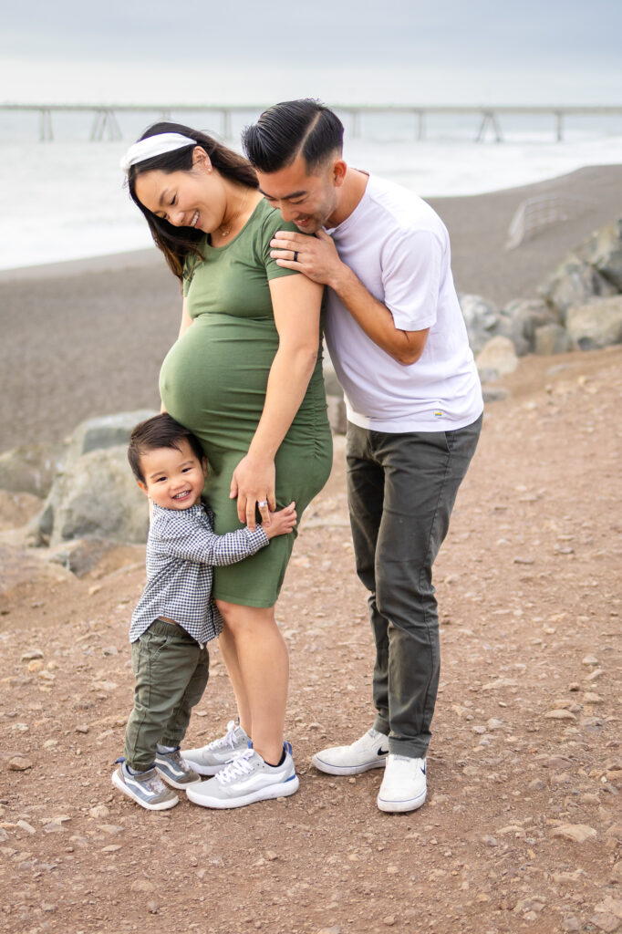 Toddler hugging his mother’s baby bump while his father leans in smiling on a breezy shoreline during Bay Area pregnancy photography.