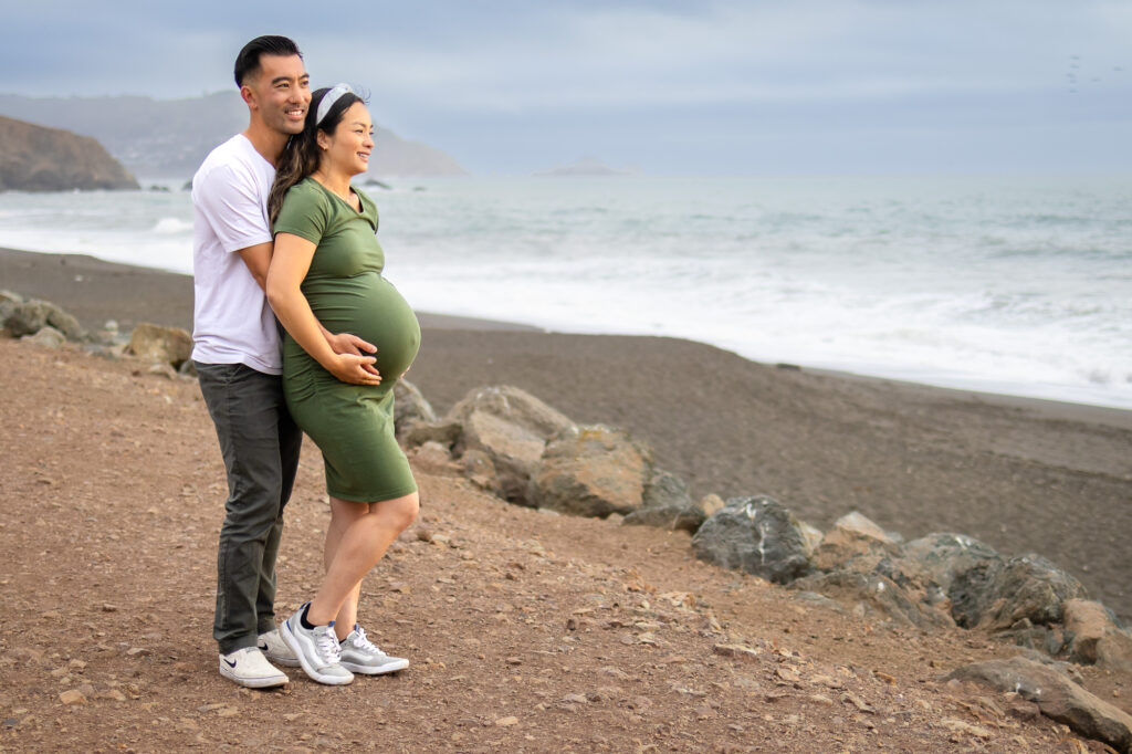 Expectant mother standing alone on a coastal path, gently holding her belly and smiling toward the camera during Pregnancy Photography in the Bay Area.