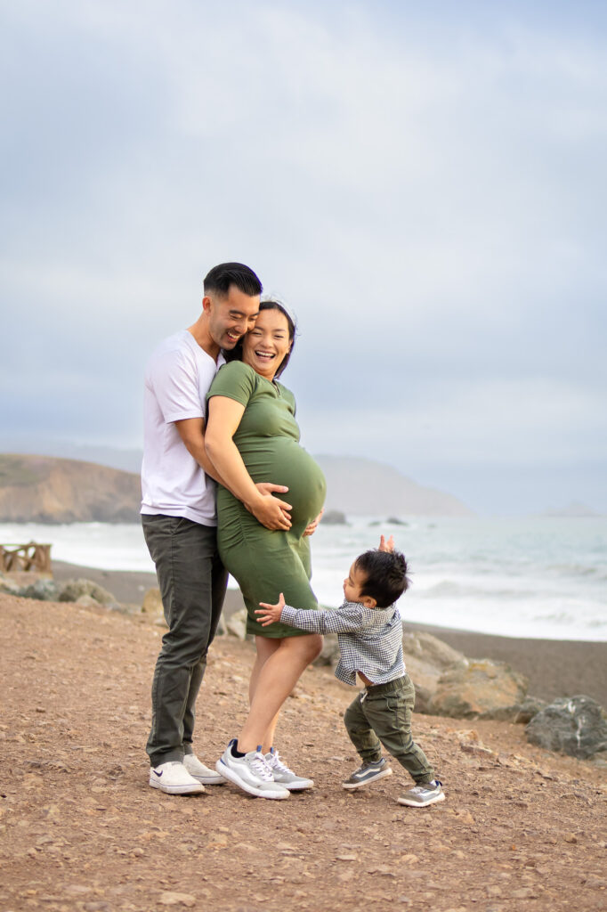 Expectant parents standing together on a cliffside beach, the father holding the mother’s belly as waves roll in behind them during calm maternity photos in the Bay Area.