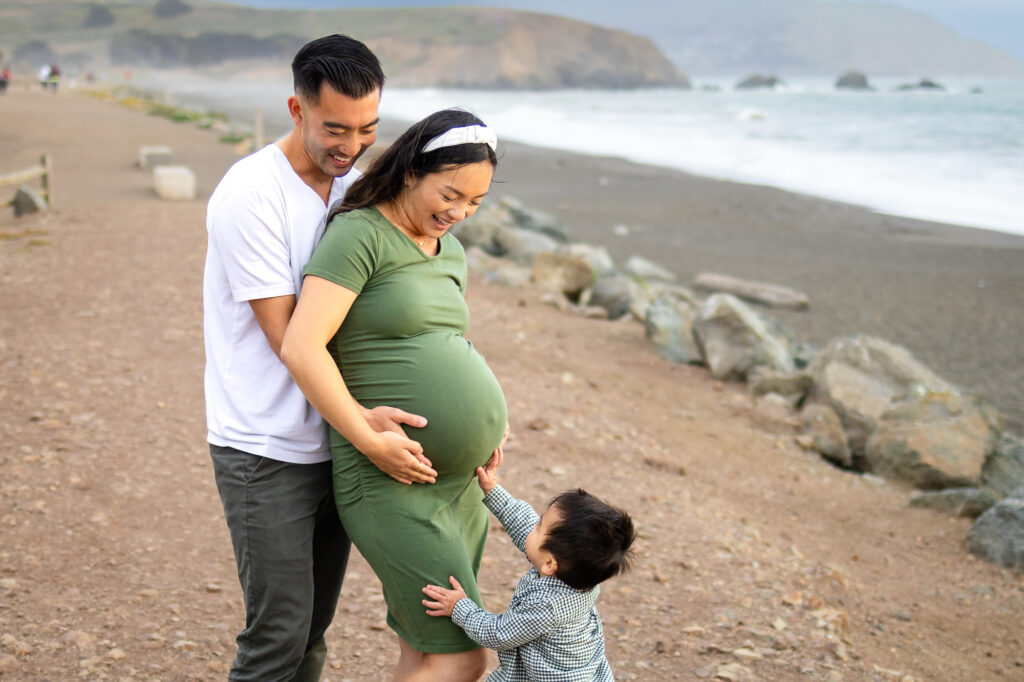 Parents laughing together as their toddler reaches up toward his mother’s belly on a seaside trail during lifestyle maternity photography in the Bay Area.