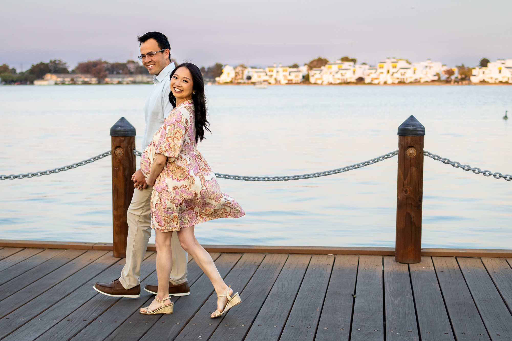 Couple walking hand in hand along a lakeside dock, the mother’s dress flowing as they smile back toward the camera during calm maternity photos in the Bay Area.