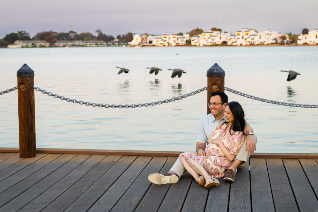 Expectant parents seated on a wooden dock by the water as birds fly overhead, sharing a quiet laugh during lifestyle maternity photography in the Bay Area.
