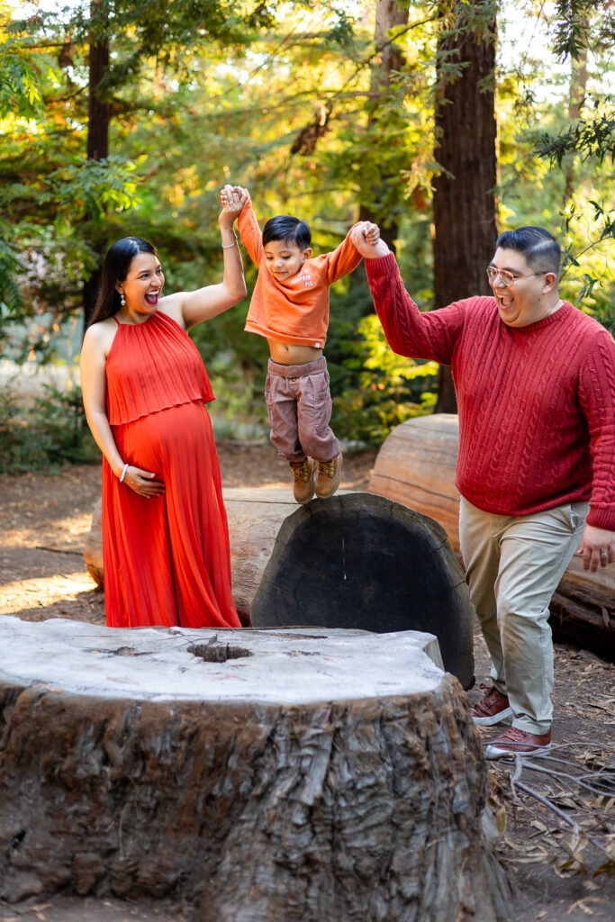 A joyful family, including a young toddler, playing among the redwoods during a maternity shoot, highlighting natural movement and a stress-free experience.