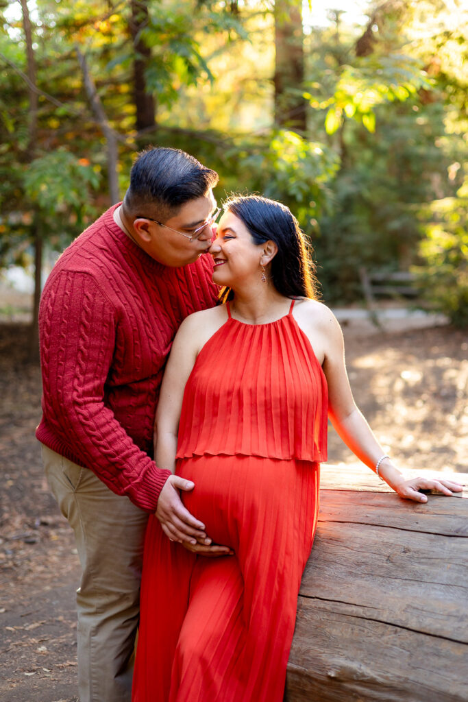 Partner standing behind a pregnant mother in a burnt orange dress, kissing her cheek and holding her belly as sunlight filters through the trees during Bay Area pregnancy photography.