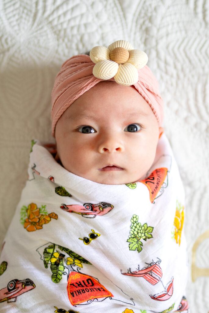 Newborn baby wrapped in a patterned swaddle and wearing a soft pink headband, photographed in natural light during an in home session.
