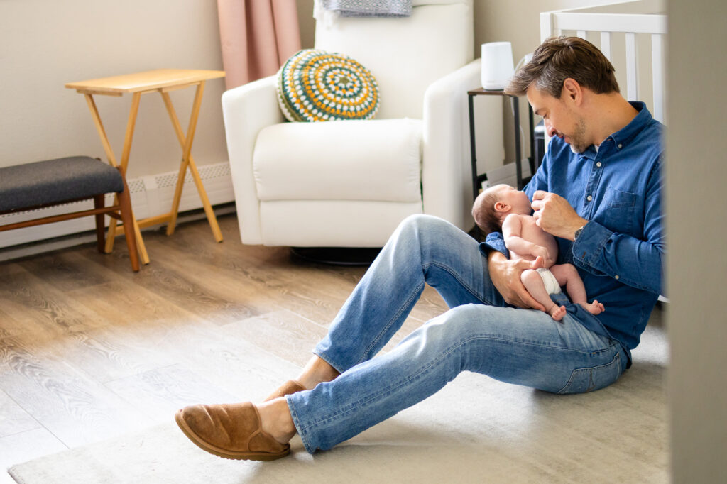 Mother and father sitting together on the nursery floor holding their newborn baby and smiling at each other during a relaxed lifestyle session.