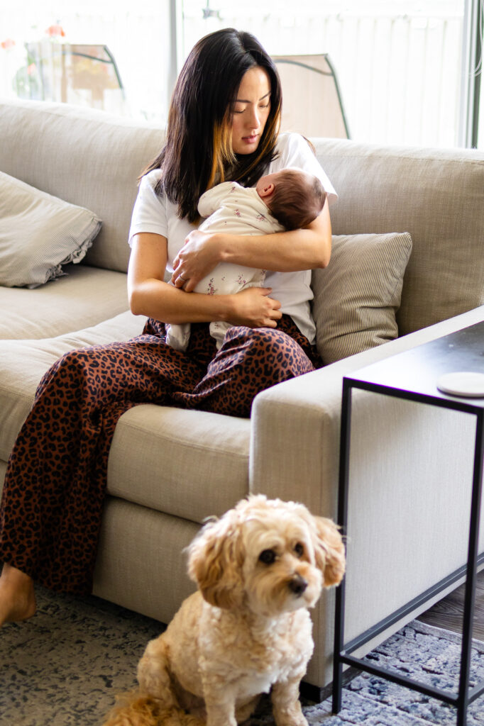 Mother sitting on a beige sofa holding her newborn close while their small dog sits on the rug in front of them in a bright living room.