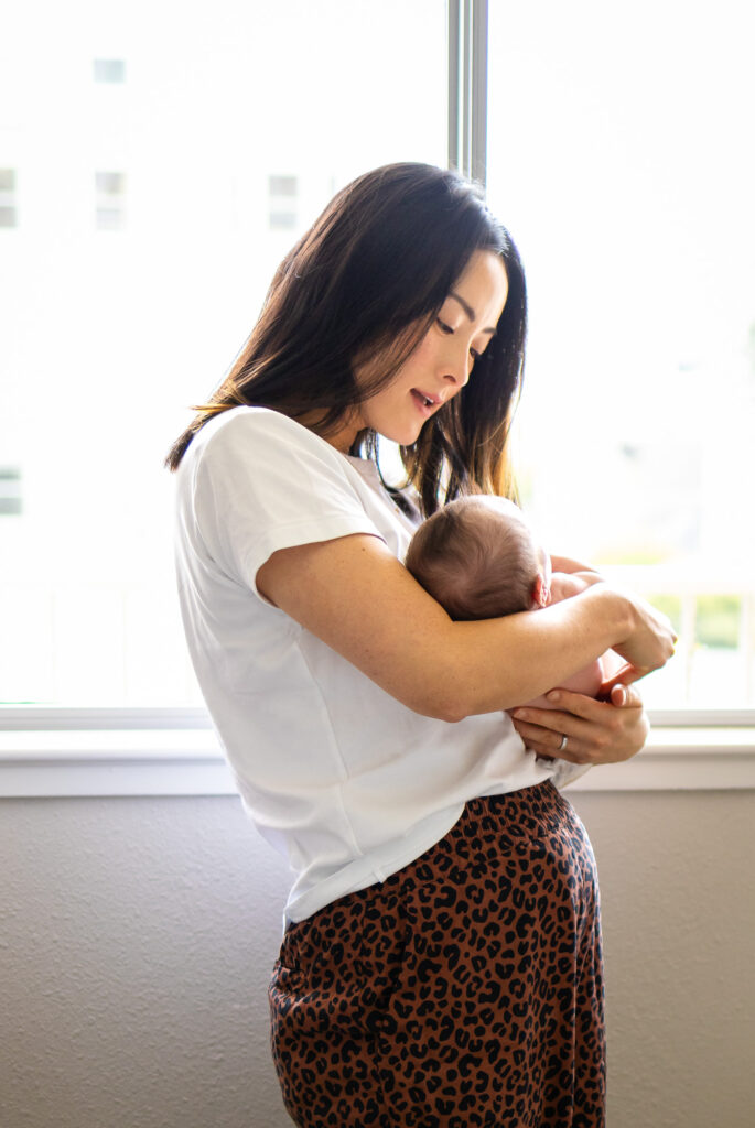 Mother standing near a bright window gently cradling her newborn against her chest in soft natural light.