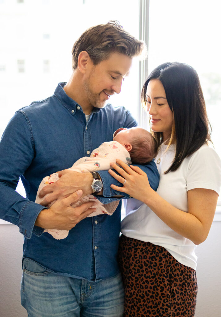 Parents standing by a window smiling down at their swaddled newborn while holding the baby together.