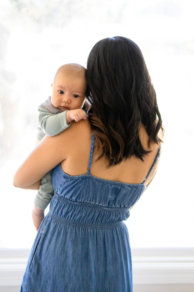 Mother holding her baby over her shoulder while the baby looks toward the camera in soft window light.