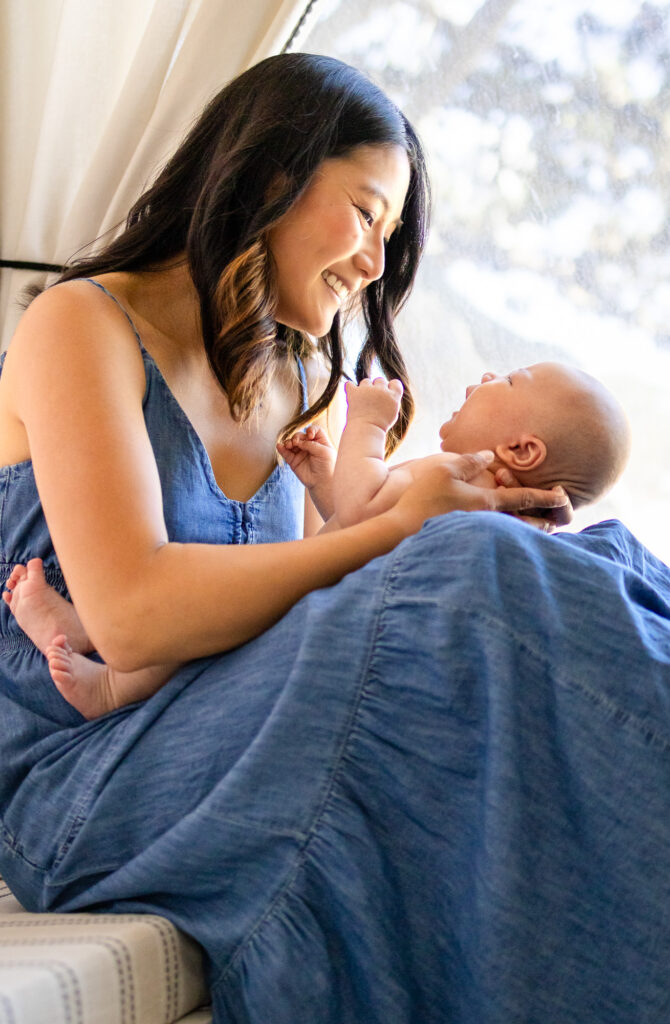 Mother smiling down at her baby while sitting by a window in a soft blue dress.