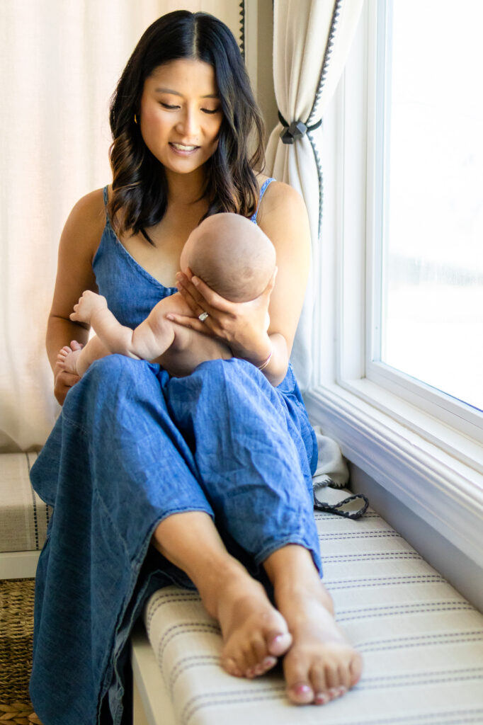 Mother sitting on a window seat holding her baby upright while looking down with a calm expression.