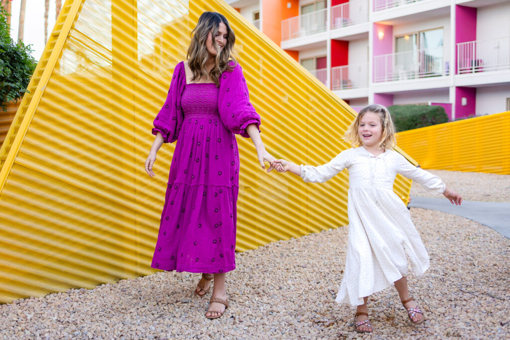 Mother holding daughter’s hand while walking along bright yellow wall during Motherhood mini sessions Peninsula lifestyle photography session