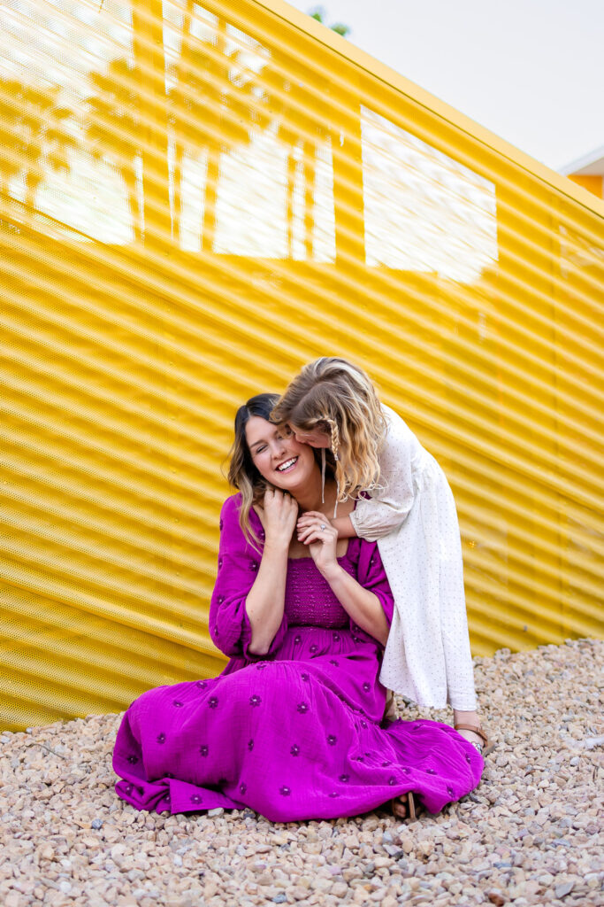 Daughter hugging mom from behind while sitting on gravel during joyful Motherhood mini sessions Peninsula outdoor portrait